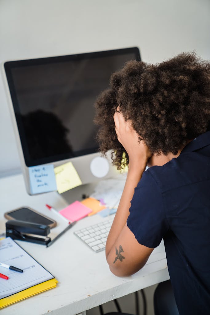JuicyCRM: Your Business Command Centre | African American woman overwhelmed at work, sitting at desk with head in hands.